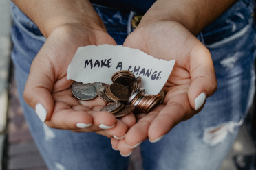 A person holds coins and a piece of paper that says "MAKE A CHANGE" in their cupped hands, with denim jeans visible in the background.