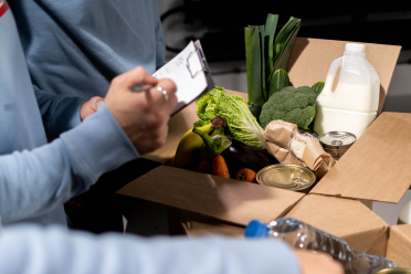 Two people check a clipboard beside an open box containing groceries including vegetables, canned food, a milk jug, and a water bottle.