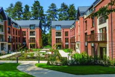 Modern apartment buildings with red brick facades and balconies surround a landscaped courtyard with pathways and greenery, set against a backdrop of tall trees.