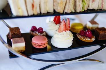 A tiered tray with assorted pastries, including a macaron, whipped cream desserts with strawberries, and square cakes, is set on a white tablecloth.