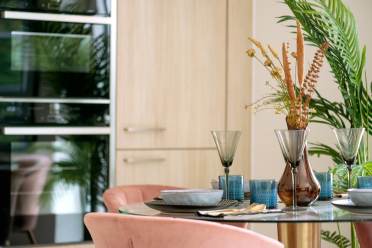 Dining table set with dishes and glasses, featuring a vase with dried flowers. Pink chairs surround the table. Wooden cabinets and green plants are in the background.