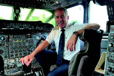 Pilot sitting in the cockpit of an aircraft, surrounded by controls and dials, wearing a uniform with captain's stripes.