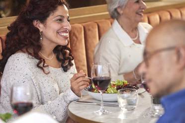 Two women sit at a restaurant table enjoying a meal and conversation. One holds a glass of red wine. A man is in the foreground, partially facing away, looking at a menu.
