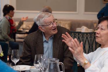 An elderly man in a checkered shirt and blazer listens attentively to a woman speaking, seated at a restaurant table with glasses and a jug. Other patrons are visible in the background.