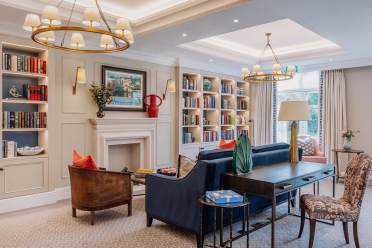 Library Elegantly decorated living room with a blue sofa, armchairs, bookshelves, a fireplace, and ceiling lights. A painting and vase with flowers adorn the mantle. Large windows provide natural light.