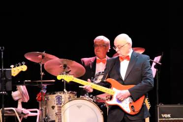 An older man in a tuxedo plays an electric guitar on stage, while another man in a suit plays drums behind him. Both are under stage lighting with musical equipment visible.