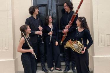Five musicians dressed in black stand in front of a door, holding a flute, clarinet, oboe, bassoon, and French horn, and looking at each other.