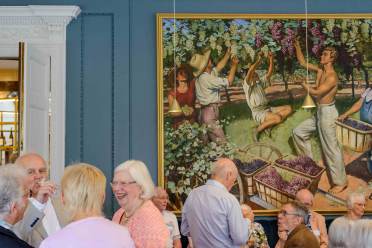 A group of elderly people socializes in a room with blue walls, beneath a large painting of workers harvesting grapes into baskets.