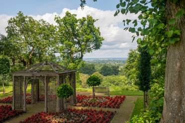 A wooden gazebo stands in a landscaped garden with red flowers, surrounded by trees, a bench, and a view of distant hills under a partly cloudy sky.