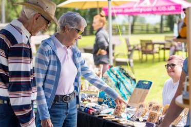 Two older adults browse a market stall displaying crafts and artwork outdoors while a vendor sits behind the table on a sunny day.
