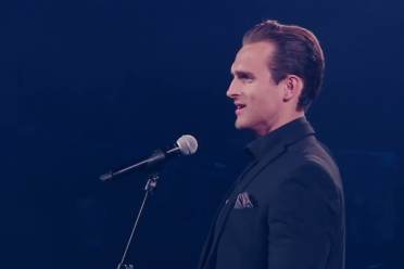 Harry Kersley A man in a dark suit stands at a microphone on stage, facing sideways, under blue lighting.