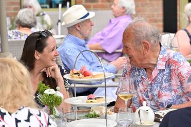 Two people sit at a table outdoors, smiling and talking, with plates of desserts and glasses of wine in front of them. Other diners are visible in the background.