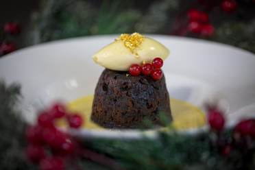 A Christmas pudding topped with a scoop of cream and red currants on a plate with custard.