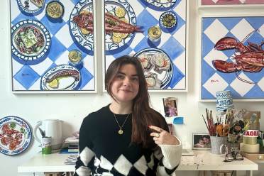 A woman sits in front of several paintings of Mediterranean-style meals and seafood, with art supplies on her desk.