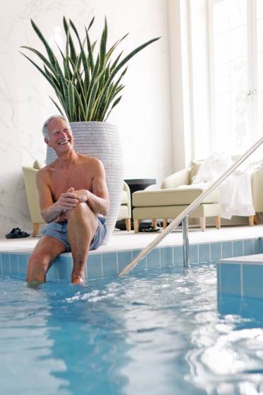 An older man in swim trunks sits at the edge of an indoor pool, smiling, with a large potted plant and chairs in the background.
