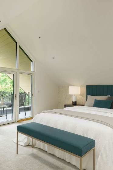 A modern bedroom with a large window and balcony. It includes a bed with a teal headboard, two nightstands with lamps, a beige armchair, and a teal bench at the foot of the bed.