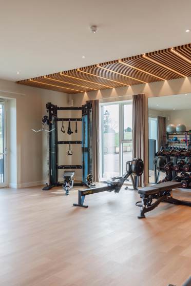 A modern gym with exercise machines, weights, and workout benches. Large windows and a mirrored wall enhance the spacious interior.