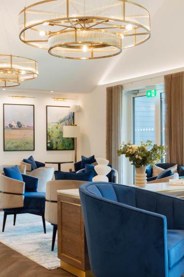 Luxurious lounge area with blue velvet chairs, round chandeliers, large windows, and framed artwork. Neutral tones with gold accents create an elegant atmosphere.