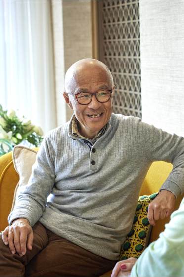 Two elderly people sitting on a yellow sofa, engaged in conversation, with light shining through sheer curtains in the background.