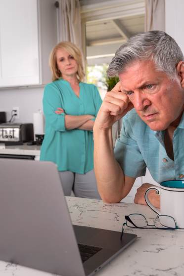 A man leans on a kitchen counter, looking at a laptop with a thoughtful expression. A woman stands behind him with crossed arms. A mug and glasses are nearby.