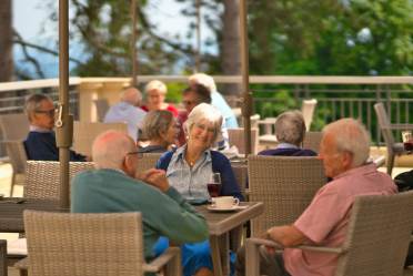 A group of older adults sit at outdoor tables on a patio, talking and drinking, with trees and a railing visible in the background.
