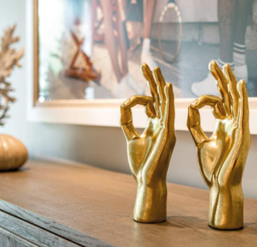 Two golden sculptures of hands making an "OK" gesture are displayed on a wooden surface in front of a framed picture.