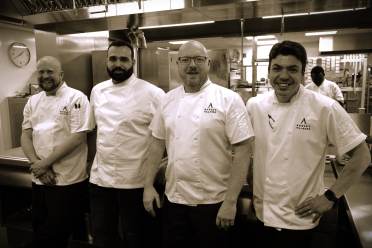 Four chefs in white uniforms stand in a commercial kitchen, smiling.