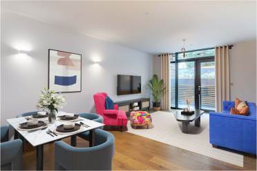Modern living room and dining area with a blue sofa, pink chair, dining table set for four, and a wall-mounted TV. Large windows with curtains provide natural light. A plant decorates the corner.
