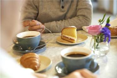 Two people sitting at a table with coffee cups, a slice of cake, and a croissant. A vase with pink and purple flowers is on the table.