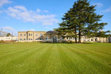 A large, historic building with a symmetrical facade stands behind a vast, well-maintained lawn and a tall tree under a clear blue sky.