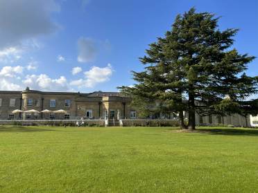 A large historic building with stone columns sits behind a spacious green lawn and a tall evergreen tree under a blue sky with scattered clouds.