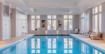 Indoor swimming pool with clear blue water, surrounded by beige tiles. Large windows let in natural light. Several lounge chairs and tables are placed along the sides.