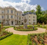 A large, white historic building with classical architecture stands behind a circular garden featuring a central stone urn and landscaped paths.