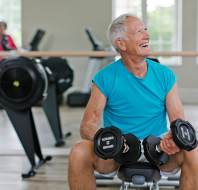 An older man wearing a blue sleeveless shirt sits on a bench holding dumbbells and smiling in a brightly lit gym, with exercise equipment in the background.
