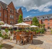 Outdoor patio area with wooden tables and chairs under umbrellas, surrounded by blooming flowers, in a courtyard between red-brick buildings on a partly cloudy day.