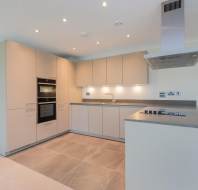 Modern kitchen with beige cabinets, built-in oven, gray countertops, electric stovetop, and under-cabinet lighting, featuring large floor tiles and a stainless steel range hood.