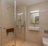 Modern accessible bathroom with beige tiles, walk-in shower with a mounted seat and grab bars, wall-mounted sink, toilet, and a rectangular mirror above the sink.