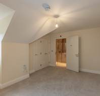 Empty beige carpeted room with sloped ceiling, closed closet doors, an open door leading to a bathroom, and a window with a blind on the left.