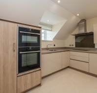 Modern kitchen with built-in double ovens, light wood and beige cabinetry, a sink under a window, and a stainless steel range hood.