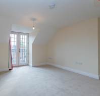 Empty, carpeted room with cream walls, sloped ceiling, and French doors leading to a small balcony; ceiling light fixture is visible.