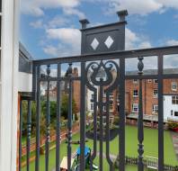 View from a balcony with ornate black railing overlooking a school playground with artificial grass and outdoor play equipment, surrounded by brick buildings under a partly cloudy sky.