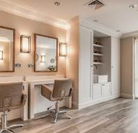 Modern salon interior with two styling stations, beige chairs, large mirrors, wall lights, built-in shelving, and floor-to-ceiling curtains allowing natural light.