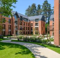 Modern brick apartment buildings with large windows and balconies, surrounded by green lawns and trees, under a clear blue sky. A curved path leads through the landscaped area.