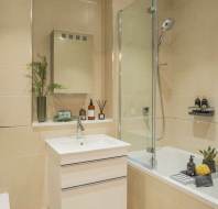 A modern bathroom with a white sink and cabinet, a bathtub with a shower, decorative plants, and toiletries. Beige tiles cover the walls and floor.
