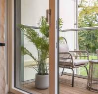 A balcony with wicker seating and a potted plant is seen through an open glass door. A small table holds a champagne bottle in an ice bucket. Trees are visible in the background.
