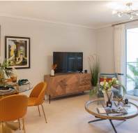 A modern living room with a round dining table, orange chairs, a TV on a wooden console, and decorative art on the walls. Large window overlooking a balcony with plants.