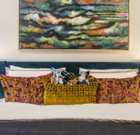 Bed with colorful pillows, a textured mustard cushion, flanked by red lamps, beneath an abstract painting with swirling colors.
