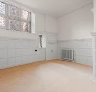 A room with white paneled walls, beige carpet, a radiator, and stained-glass windows.