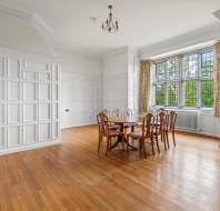 A bright dining room with a wooden table and chairs, hardwood flooring, white paneled walls, and a large window with a view of greenery.