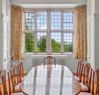 A polished wooden dining table surrounded by chairs is placed in front of a large, multi-pane window with patterned curtains. The window offers a view of greenery and a clear sky outside.